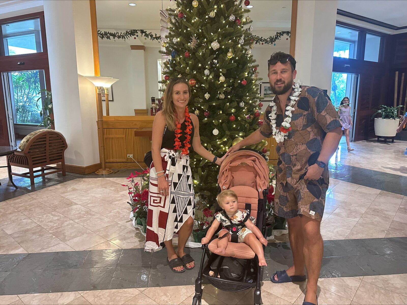 Man and woman stand in front of a christmas tree with baby in a pushchair